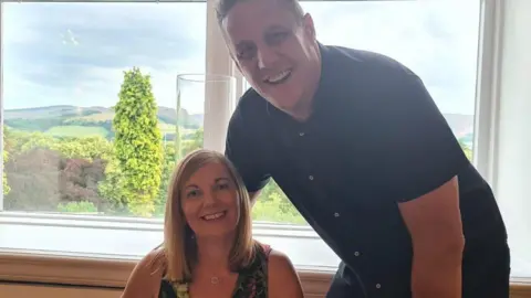 Gary Gray A woman, Anna, sits down next to a man, Gary. Anna has shoulder length blonde hair and wears a tropical patterned top. Gary has a dark shirt on. They are both smiling into the camera.