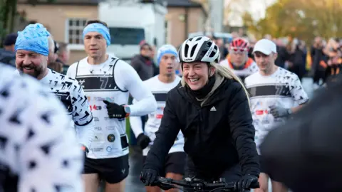 PA Media Dame Laura, smiling in a black-and-white cycling helmet, rides a bike, with Sir Jason, Sinfield and other support runners alongside and behind her. Well-wishers surround the group.