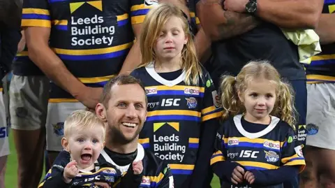 PA Media Rob Burrow, holding his son with his daughters behind him, after playing in a testimonial match at Leeds in January 2020