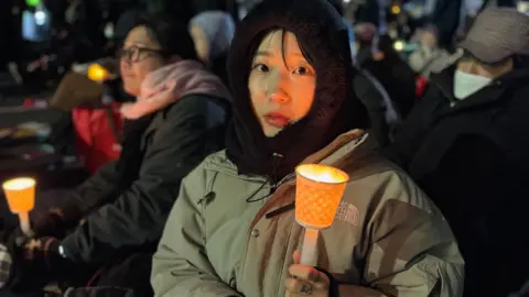 A woman looks at the camera whilst she holds a candle. She's wearing a heavy green warm coat and a black snood. Her face looks concerned, almost anxious. in the background are several other people, all wrapped up for cold weather - some also carry candles.