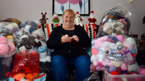 Rachel Williamson Rachel Williamson smiling on a sofa, holding knitting needles, surrounded by balls of wool