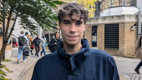 Dylan with brown hair, standing in a street, wearing a coat and smiling