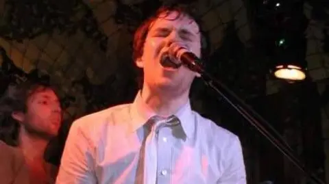 Chris Steele Brandon Flowers passionately singing into a microphone. He is wearing a white shirt and white tie, and stage lights illuminate the right side of his face with a pink hue. A bandmate looks on in the background.