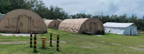 Photo shows three round beige tents and a white tend on green grass. In the background there are trees