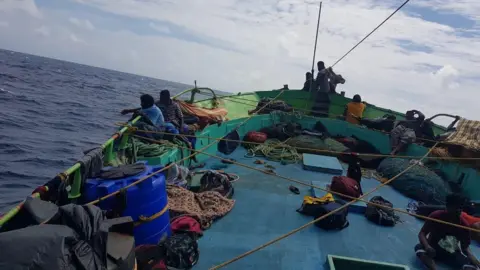 A group of people are sat on the hull of a boat which is at sea. They are surrounded by ropes and bags