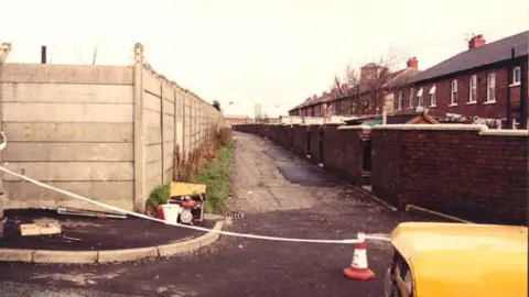 GMP Police cordon across alleyway, between the backs of terraced housing and fences.