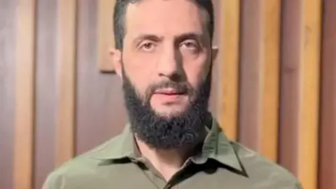 Reuters Abu Mohammed al-Jawlani looks directly at the camera in this screenshot of a video. He's dressed in a green military shirt and has a dark beard.