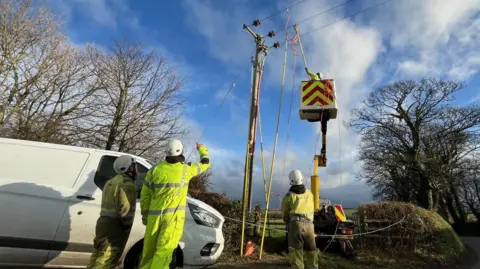 Scottish Power three workers in high viz clothing standing beside a white van parked beside a telegraph poll which has a cherry picker beside it and another worker reaching up towards a the wires