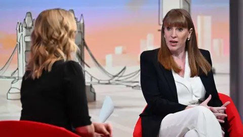 Angela Rayner, who has long red hair with a fringe and is wearing a black blazer with a white jumpsuit, sits on a chair as she speaks to the BBC's Laura Kuenssberg