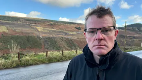 A man with short, dark hair and wearing glasses and a black coat is to the right of frame. Behind him is a road and in the distance is a hillside which is the site of a coal tip which has been made safe. Aside from the green, yellow and red colours of the grass and bracken on the hillside, there are also patches of black which is the coal tip.