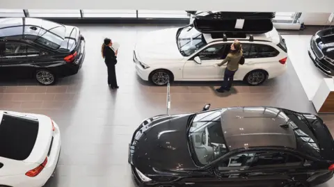 Getty Images Overhead view of a woman looking at a car in a showroom with a dealer standing nearby