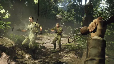 MachineGames Screenshot of a jungle scene taken from a first-person perspective shows the player's hand holding the handle of a bullwhip. The end of it can be seen striking one of two enemy characters dressed in combat fatigues about 10 feet away from the player. The one who's just been hit is recoiling and a pistol can be seen falling out of his hand. The other looks on with a panicked expression as he readies his own weapon.