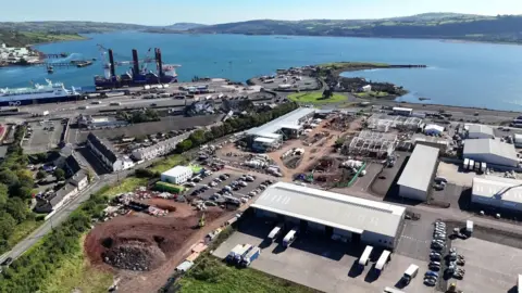 A drone image of a construction site at the harbour. There are large grey industrial sheds, an area that is being dug out and the sea is along the coast.