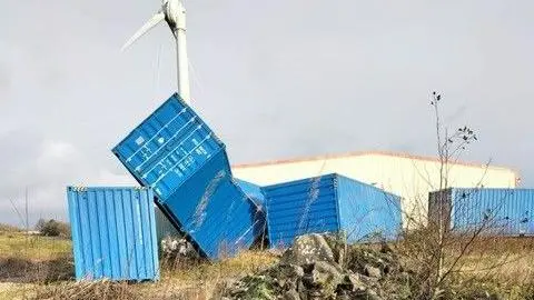 About four blue shipping containers can be seen on the ground, including one which has ended up diagonal and partially resting on a container next to it. A large building can be seen behind them.