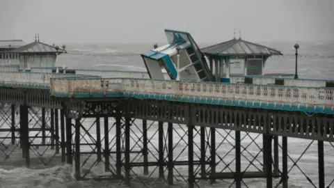 An overturned blue and white hut leans against another hut on Llandudno Pier, with large waves visible all around. Two other huts can be seen further along the pier. 