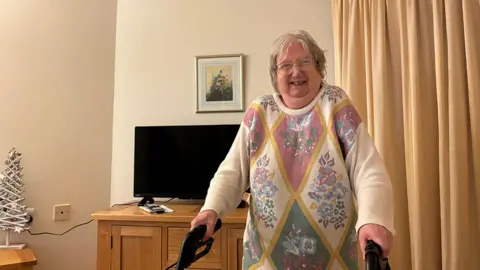 Picture of Carole taken in her new flat in Folkestone - she is holding onto a walking frame in her living room - there is a TV behind her and a small artificial Christmas tree can be seen on the far left of the picture. Carole is wearing a sweater with floral patterns on it and she is smiling broadly. 