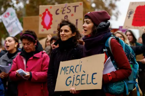 Reuters A group of women stand outside, holding placards. One woman at the front holds a cardboard sign that reads "Merci Gisèle"