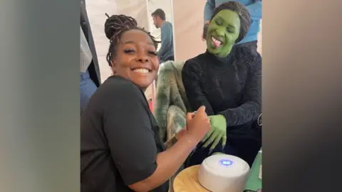 Shea Osei Shea Osei has her hair tied up and is smiling at the camera. She is wearing all black and is sat doing Cynthia Erivo's nails. Cynthia Erivo is also wearing black and has her body and face painted green. She has her eyes closed and is sticking her tongue out to the camera. The pair are sat around a wooden table with a gel lamp on it.