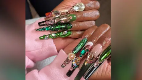 Shea Osei A close up of Cynthia Erivo's hands with her hands clasped shut and her nails on her palms. The sleeves of a pink hoodie can be seen. Eight nails can be seen each with green and pink designs. Some nails have a chrome metallic design on whereas others are encrusted with jewels and gems, making the design 3D. 