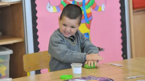 Newspics Kyran sits in a school classroom wearing a grey V-neck jumper which has a yellow band around the neck line. He has dark brown hair and brown eyes and his face is scrunched up as he molds some green play dough on the school desk. A small plastic tub bearing his name sits in front of him, with a green lid beside it. Kyran is sitting on a wooden school chair with a large pink classroom display and storage visible behind him. The display shows the lower half of a clowns body, holding a piece of sting which is likely attached to a balloon. 