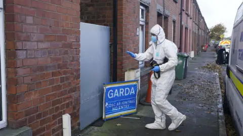 PA Media A forensic officer dressed in a full white boiler suit carries a large camera towards a sheeted steel gate, marking the entrance to an alleyway. A small blue pop-up sign sits on the ground next to him which reads "Garda". Below that writing in Irish reads "Cosc ar iontrail" followed by the English translation of "no entry". A Garda van is parked behind the forensic officer and in the background are some wheelie bins and red bricked terraced houses.