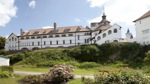 An enormous white and terracotta abbey sits on a small hill above he village on Caldey Island. It was designed by Cardiff architect John Coates Carter under the direction of Abbot Aelred Carlisle and has a distinct style. It is surrounded by a small stone wall, grass and plants. There is a single lane path or small road that lies in front of it. 