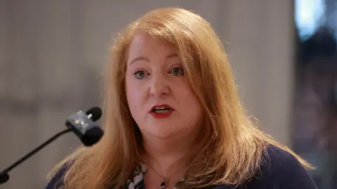 PA Media Justice Minister and Alliance leader Naomi Long has strawberry blonde and wearing a purple cardigan and speaking into a microphone.