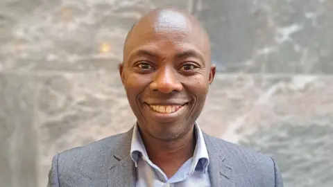 head and shoulders of man, looking at camera, bald and clean shaven, smiling, wearing grey suit