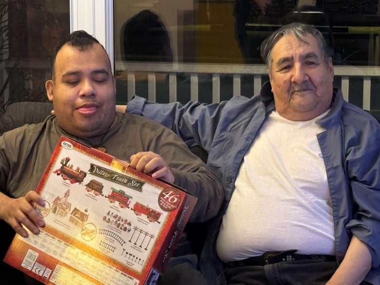 Two men sit together on a couch. One holds a box containing a toy train set. 