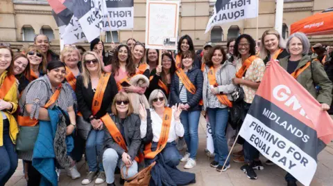 GMB Union A large group of women outside a sandstone building. All those pictured are wearing orange sashes and a woman at the front holds a large flag which says "GMB Midlands Fighting For Equal Pay". Several other women at the back hold similar flags. Two of the women are crouching down at the front and all are smiling.