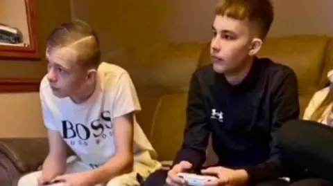 Family handout Mason Rist and Max Dixon are pictured sitting on a sofa holding PlayStation controllers and looking at a screen