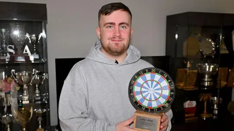 Pinpeg Luke Littler with brown hair and beard wearing a grey hoodie holding a trophy in the shape of a darts board with trophy cabinets either side of him filled with trophies 