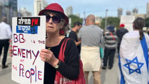 Siviona, a woman wearing a black top, a red hat and a red tote bag, looks at the camera. She holds a placard that says 'Bibi is the enemy of the people'. Behind her is a person facing away, wearing an Israeli flag as a cape.