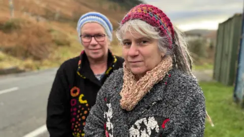 Two women are stood looking into the camera lens. The woman to the left is wearing glasses, a black cardigan with colourful circles and a stripy blue hat. The woman to the right has grey dreadlocks, tied back in a pink knitted headband. She is wearing a grey coat and a peach scarf. They both stand to the side of a road. 