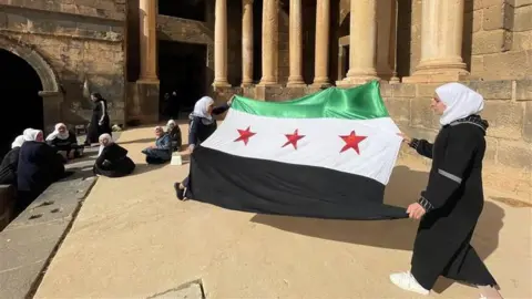 Women watch as the Syrian flag used by the opposition with a green stripe, a white stripe with three red stars and a black stripe is unfurled