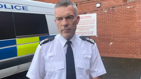 PA Media A middle-aged man with short grey hair wearing a white shirt with black epaulettes featuring a crown on the shoulders and a black tie. A police car his behind him,