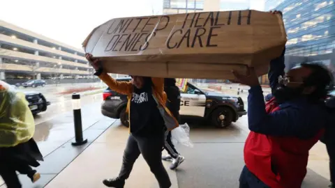 Getty Images Two people in the midst of a protest holding a coffin which says "united health denies care" on it