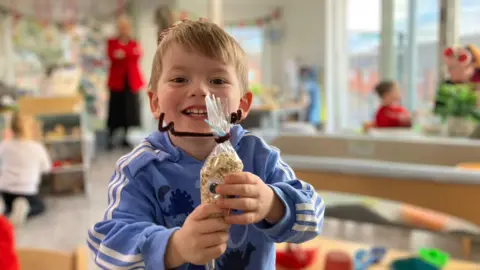 Vanessa Clarke / BBC A child smiles at the camera as he holds up a craft he has made