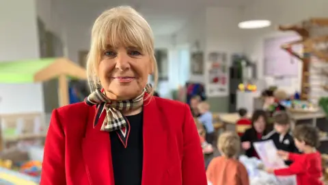 Vanessa Clarke / BBC Nicola Fleury is smiling at the camera wearing a red blazer and a neck scarf. She has blonde hair.