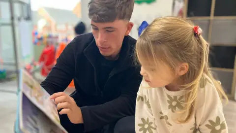 Vanessa Clarke / BBC An early years staff member reads to a child inside a nursery classroom