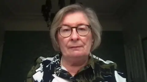 A picture of Andrea Nelson, who has short grey hair. She is wearing thick dark rimmed glasses, a pearl necklace and a top with olive green, black and white floral patterns on it. 