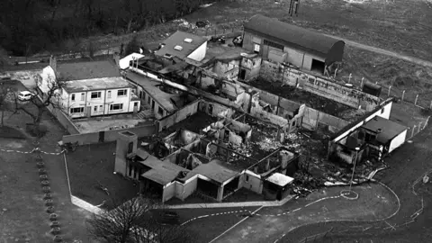 Pacemaker Black and white aerial image of the bombed La Mon hotel, the brick building is covered in black with the roof almost completely gone.  with parts with most of the building frame collapsed, debris lies on the floor.