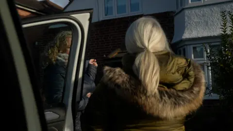 Two women stand on a driveway, their images obscured. Both are wearing thick winder padded coats, one has shoulder-length curly blonde hair and is looking towards the other woman, with long blonde hair tied back. The house can be seen in the background and in the foreground is a car door.