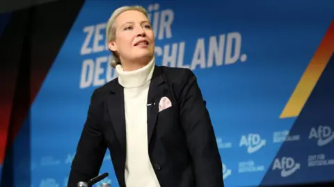 Maryam Majd/Getty Images A woman wearing a black jacked and white sweater talks to an audience of supporters from a stage in Germany