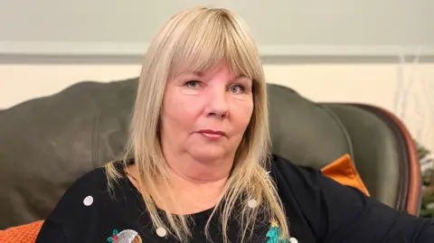 A picture of Stella McLoughlin, she has long blonde hair with a fringe and is wearing a dark jumper with a christmas tree and christmas pudding on it. She is sitting on a dark green sofa with orange cushions. 