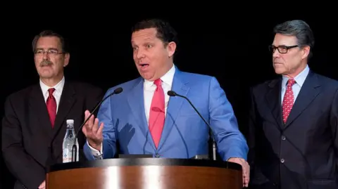 Getty Images The lawyer Tony Buzbee, wearing a light blue suit, stands at a podium during a press conference, as former Texas governor Rick Perry watches from his left hand side