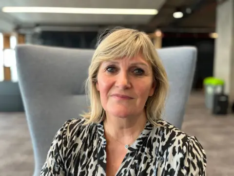 A woman with blonde hair, wearing a black-and-white top and a necklace, sits on a grey chair in an office