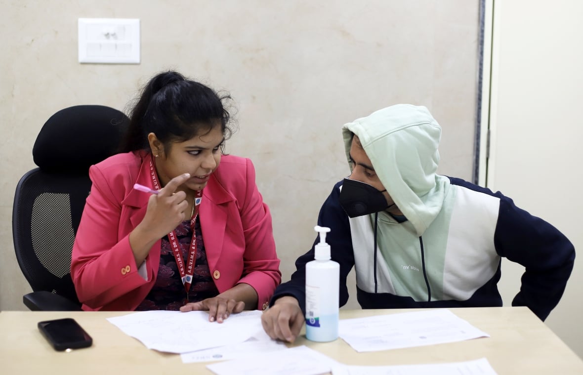 A woman and man sitting at a desk. She is filling out papers and talking to the man, who is wearing a face mask.