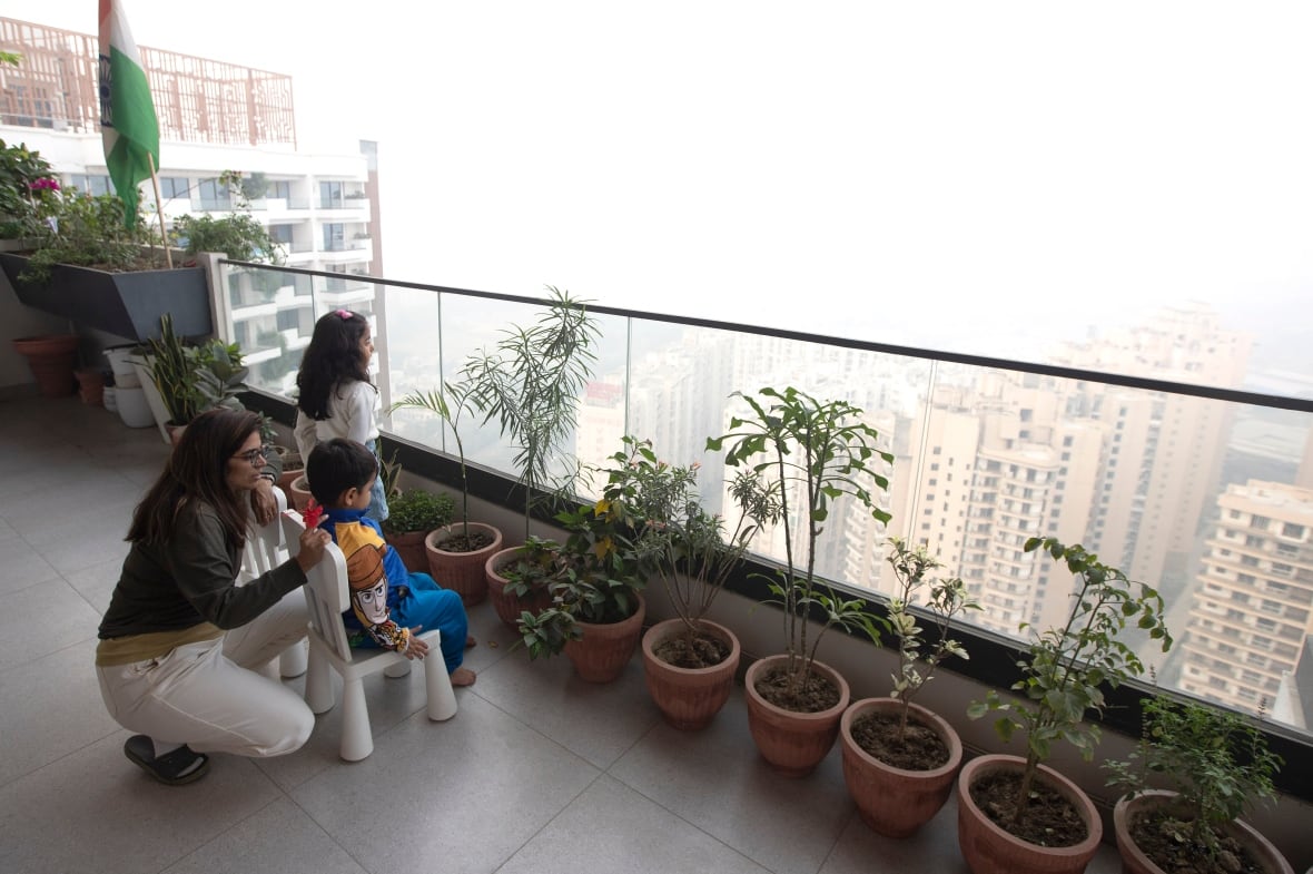 A woman crouches beside two children sitting on small chairs on a high-rise balcony. Beyond the balcony’s clear railing, heavy smog almost obliterates the city skyline. 