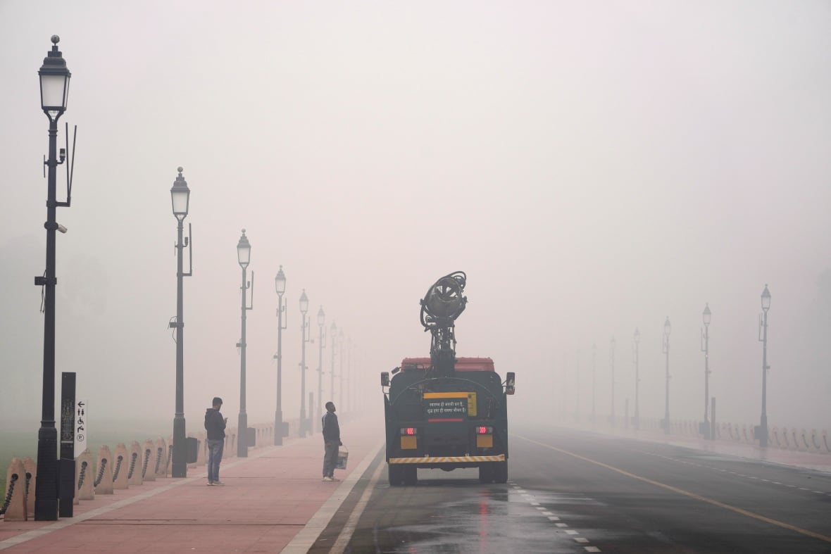 A truck drives through heavy smog along a nearly empty street. Two people stand near it on a sidewalk. 